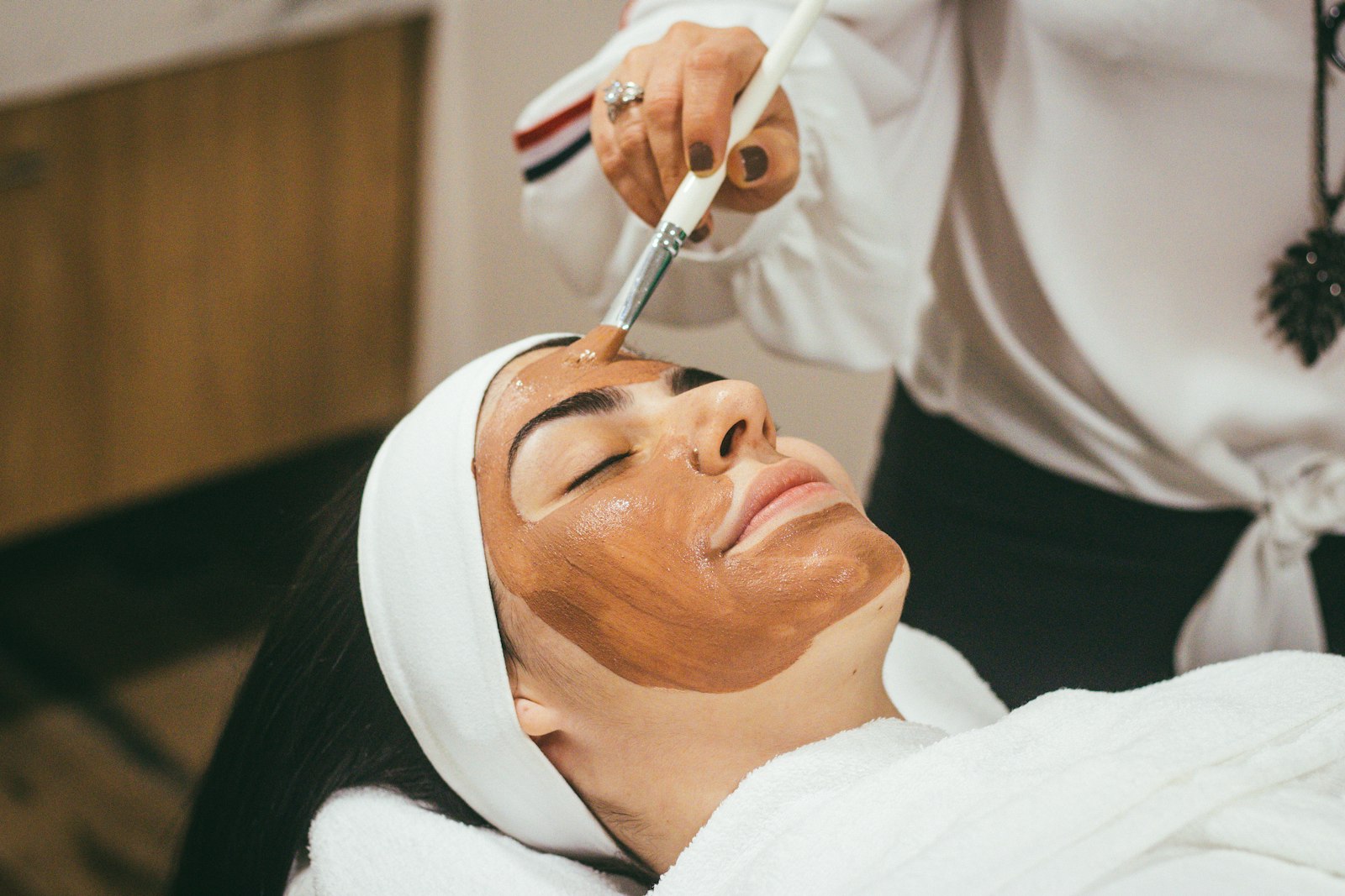 An esthetician applying a clay facial mask to a relaxed client